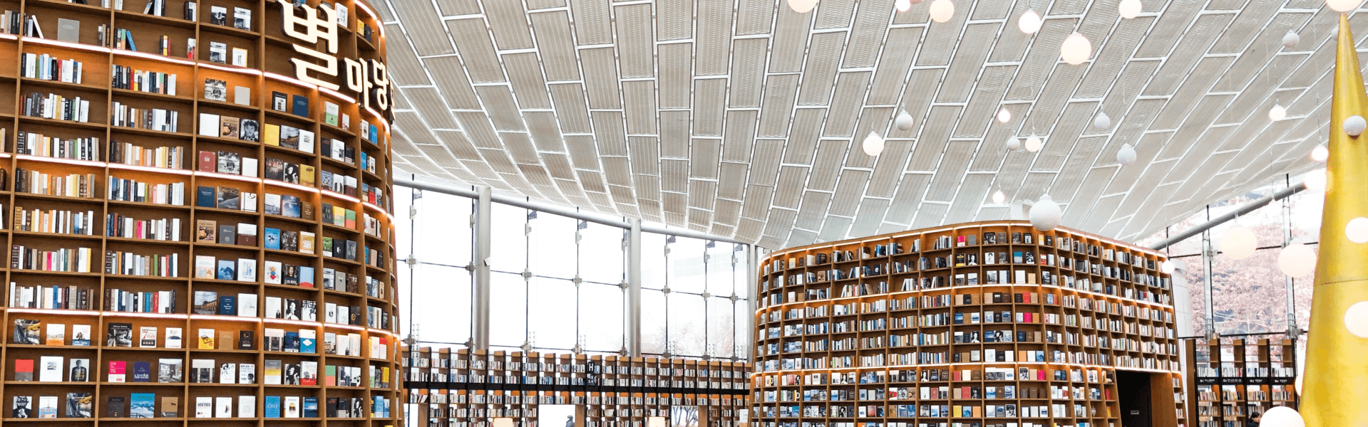 Interior of a modern library with tall windows and a white ceiling with white pendant bulbs. There are bookshelves all around the wall. On the right, a tall bookshelf forms a square of rounded edges. Its walls are slanted inwards, and it has a door on one of its sides. Another tall and slanted bookshelf is located on the left.